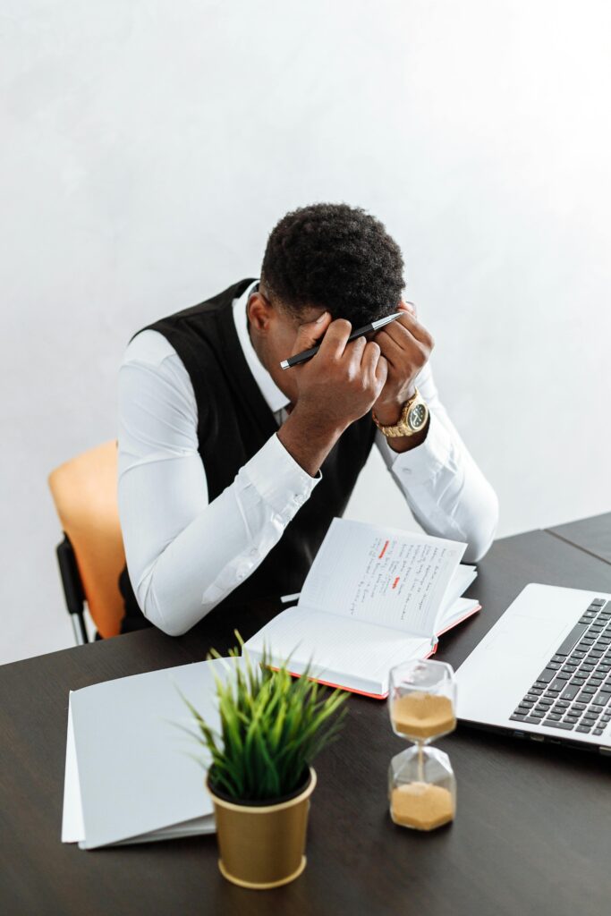 Man sitting at a desk with his head in his hands, surrounded by a laptop, notebook, and hourglass.