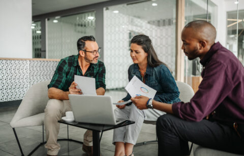 Three adults sit together reviewing documents and charts with a laptop during a discussion in an office.