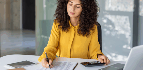 Woman in a yellow blouse using a calculator while reviewing a financial spreadsheet at her desk.