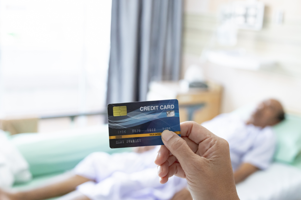 A person holding a credit card in the foreground while a patient lies in a hospital bed in the background.
