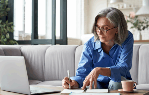 Older woman with gray hair and glasses sitting on a couch, reviewing paperwork and writing in a notebook while using a laptop at home.