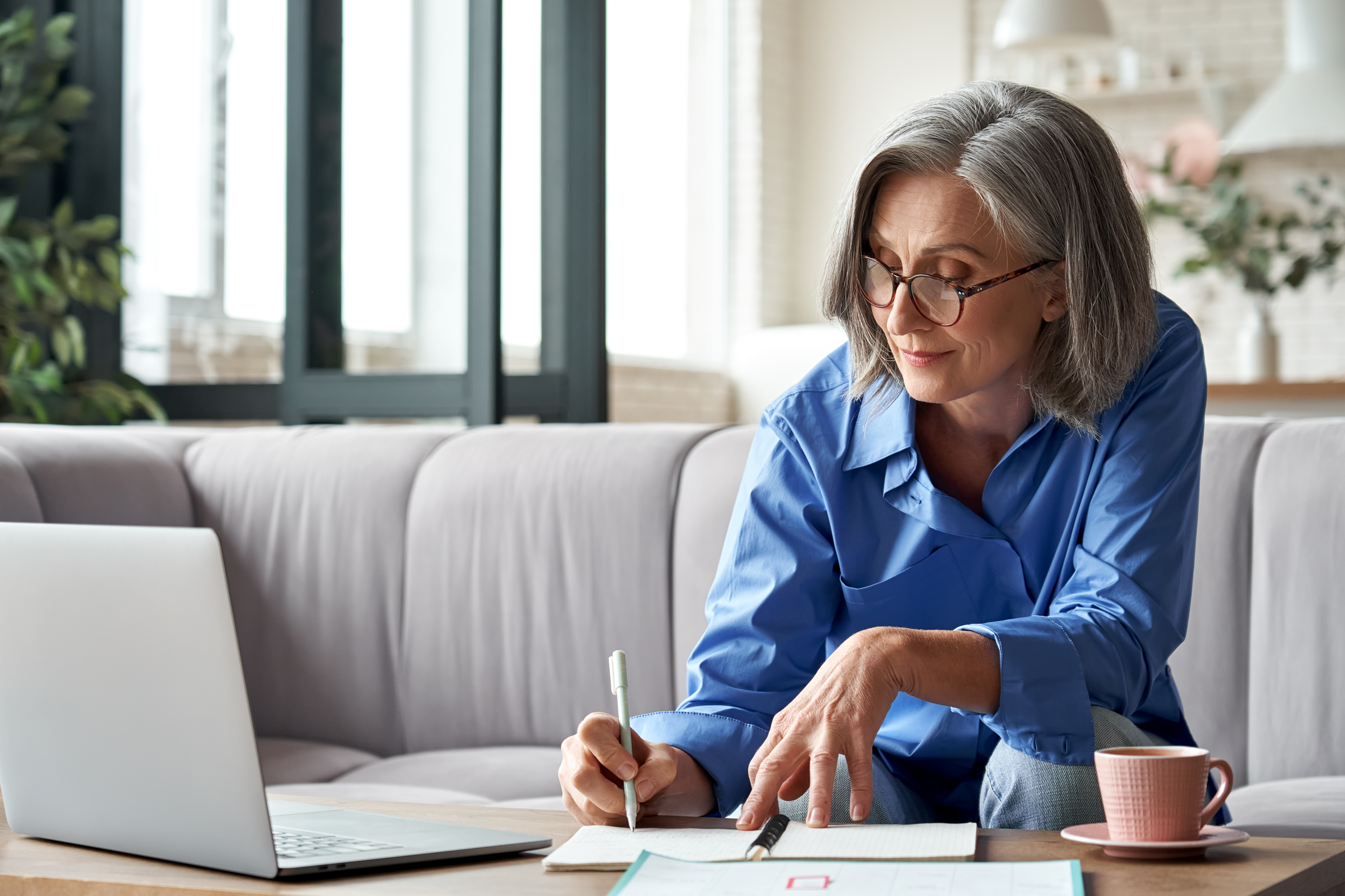 Older woman with gray hair and glasses sitting on a couch, reviewing paperwork and writing in a notebook while using a laptop at home.
