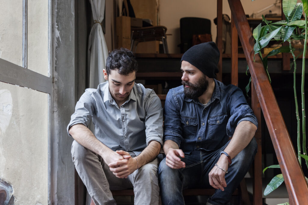 Two young men sitting on stairs indoors, one looking down with folded hands while the other looks at him and speaks.