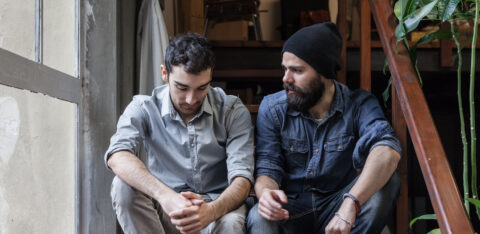 Two young men sitting on stairs indoors, one looking down with folded hands while the other looks at him and speaks.
