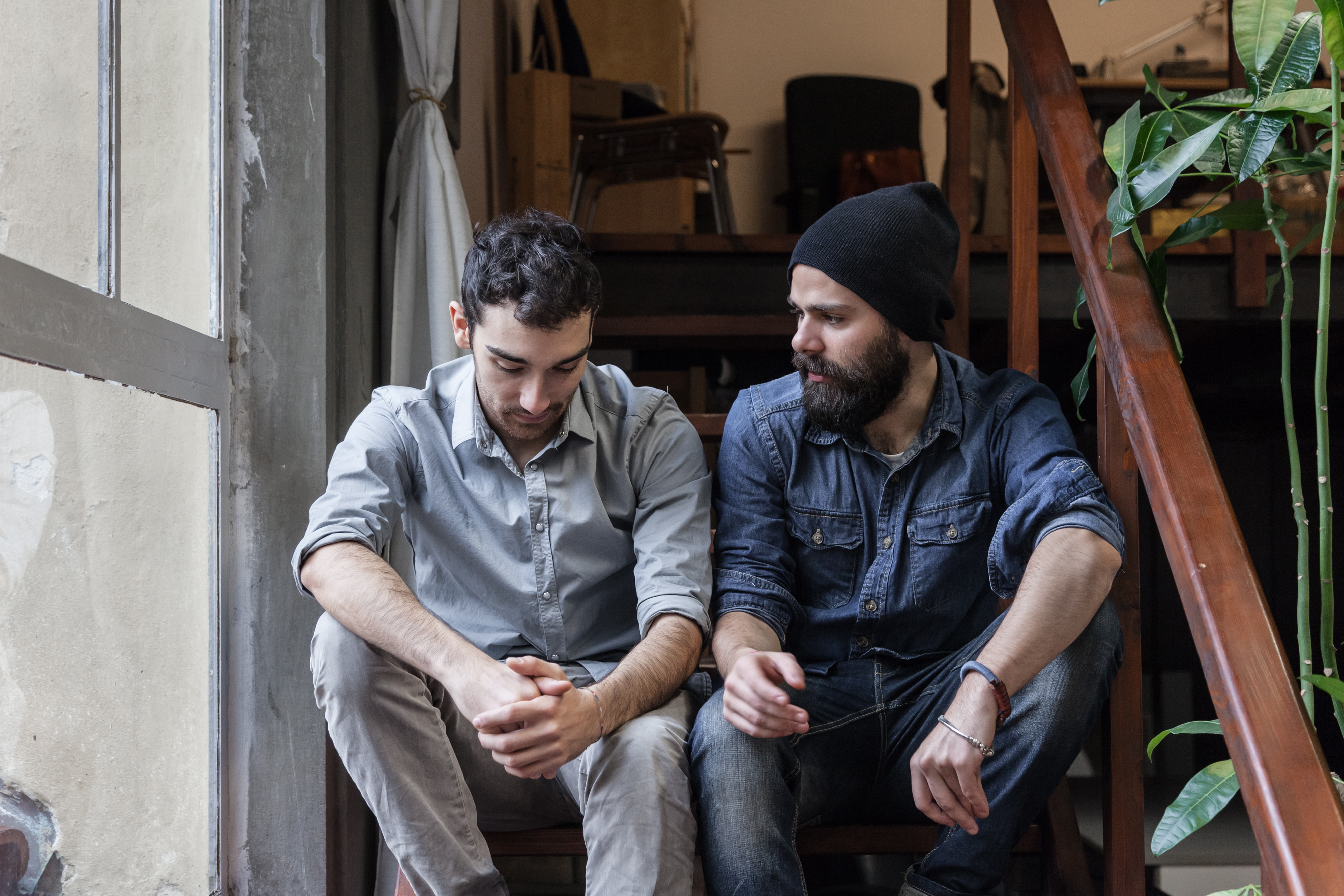 Two young men sitting on stairs indoors, one looking down with folded hands while the other looks at him and speaks.