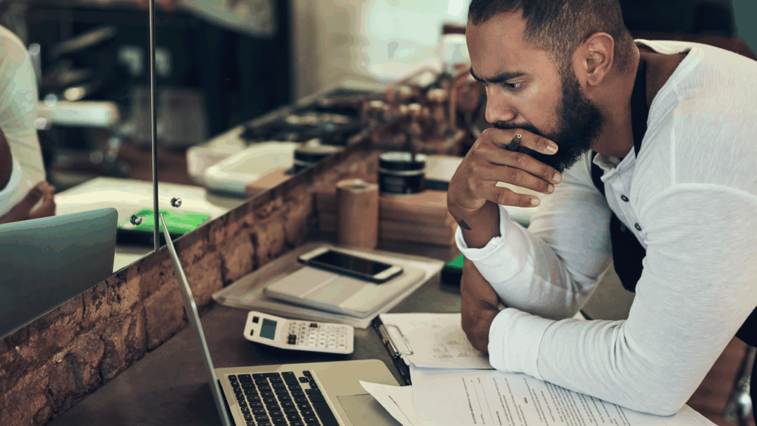 Man leaning over a counter, reviewing paperwork and using a laptop and calculator, appearing deep in thought.