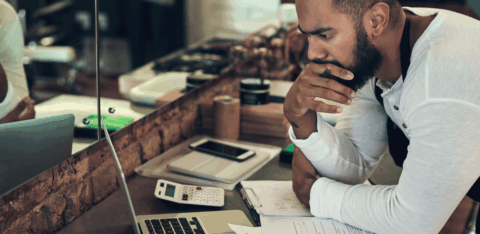 Man leaning over a counter, reviewing paperwork and using a laptop and calculator, appearing deep in thought.