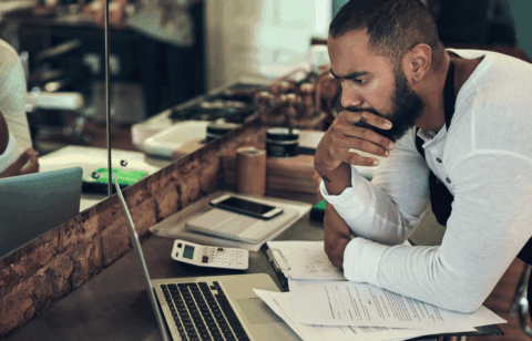 Man leaning over a counter, reviewing paperwork and using a laptop and calculator, appearing deep in thought.