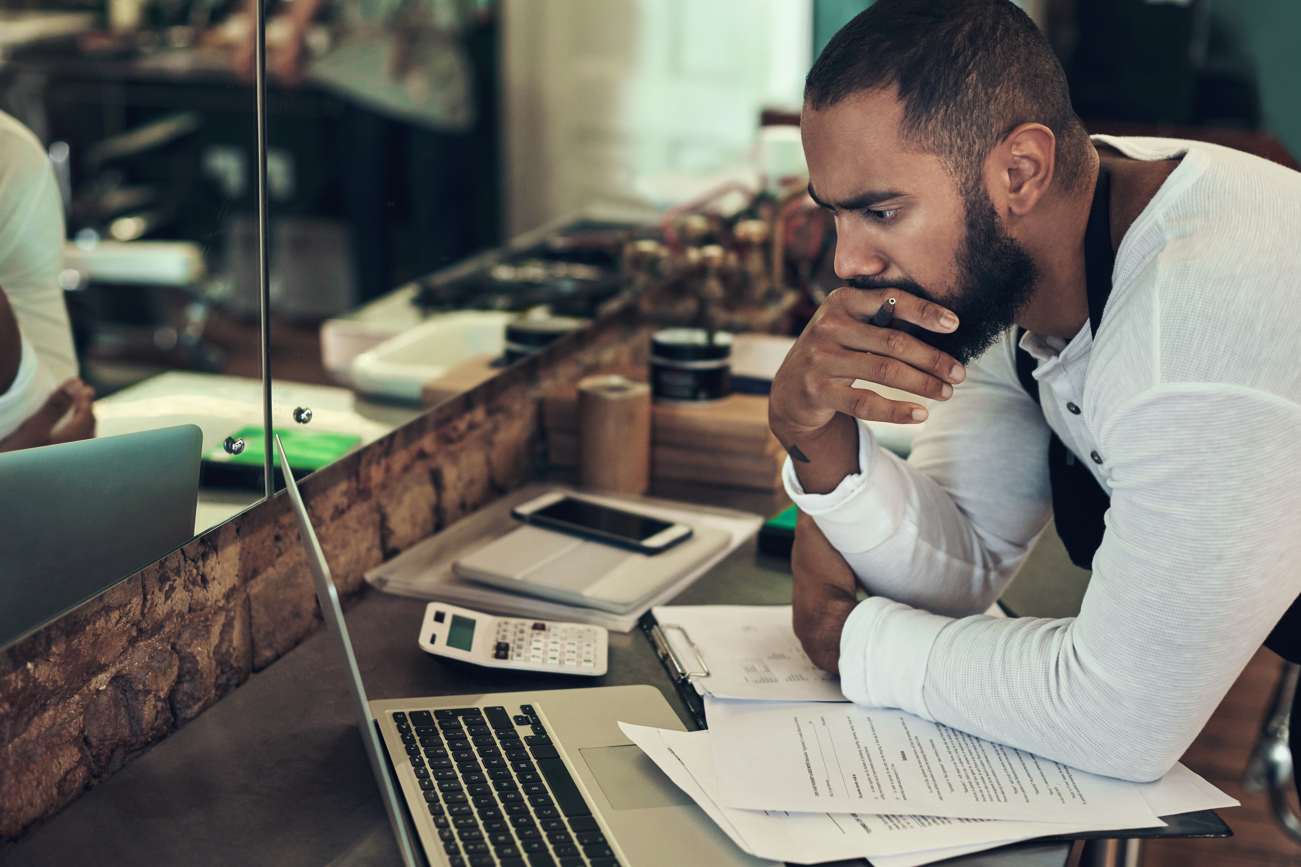 Man leaning over a counter, reviewing paperwork and using a laptop and calculator, appearing deep in thought.