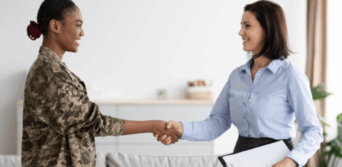 Smiling woman in military uniform shaking hands with a professional woman holding a clipboard.