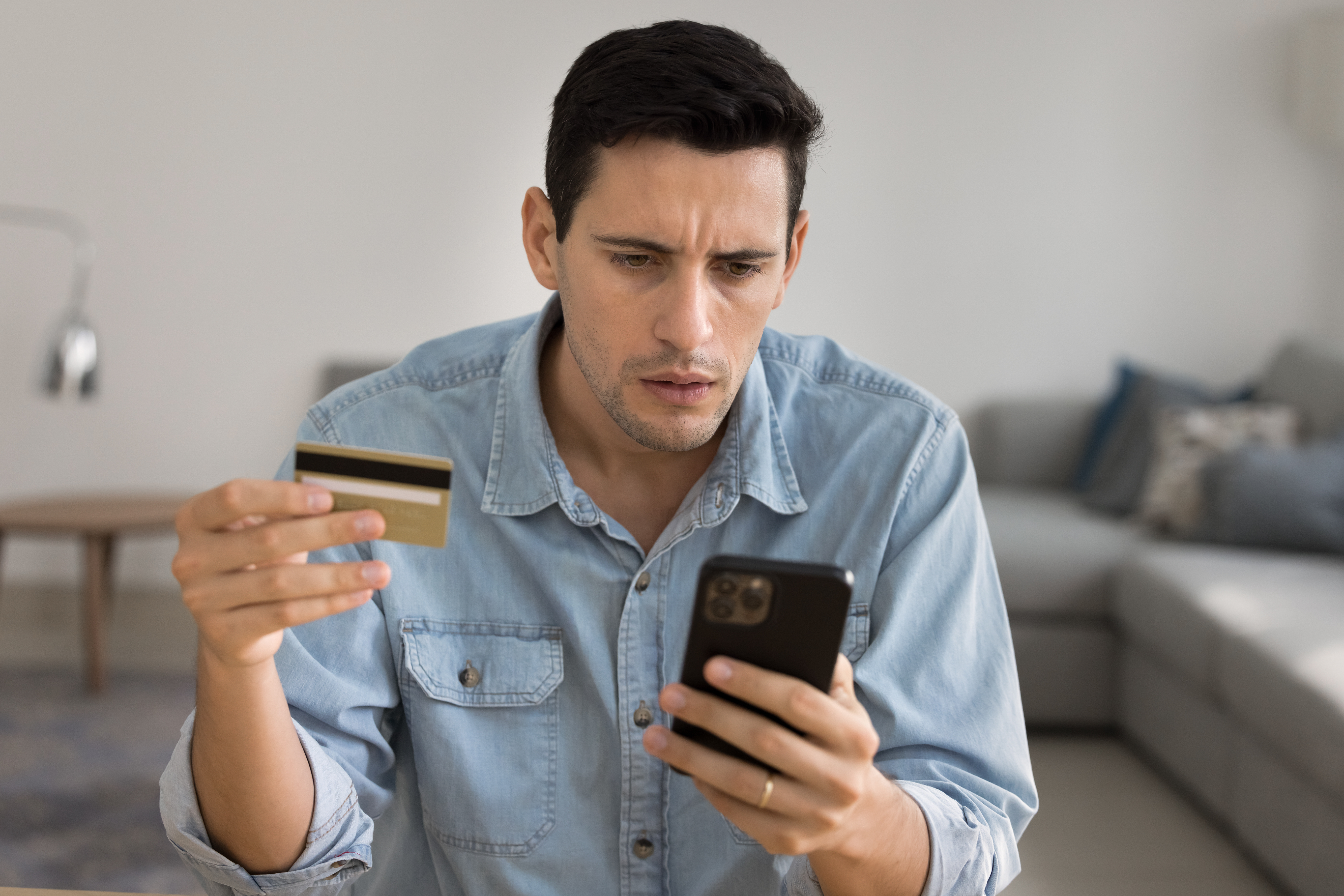Man holding a credit card and looking concerned while checking his smartphone.