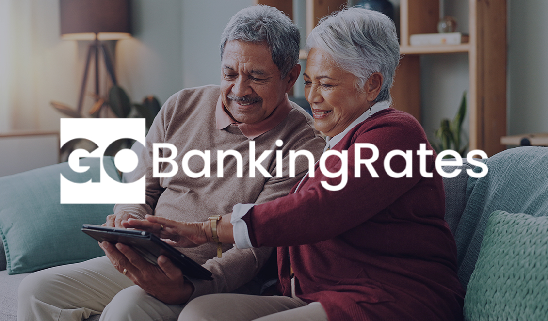 Smiling older couple enjoying time together on a couch while looking at a tablet, with the GOBankingRates logo overlaid.