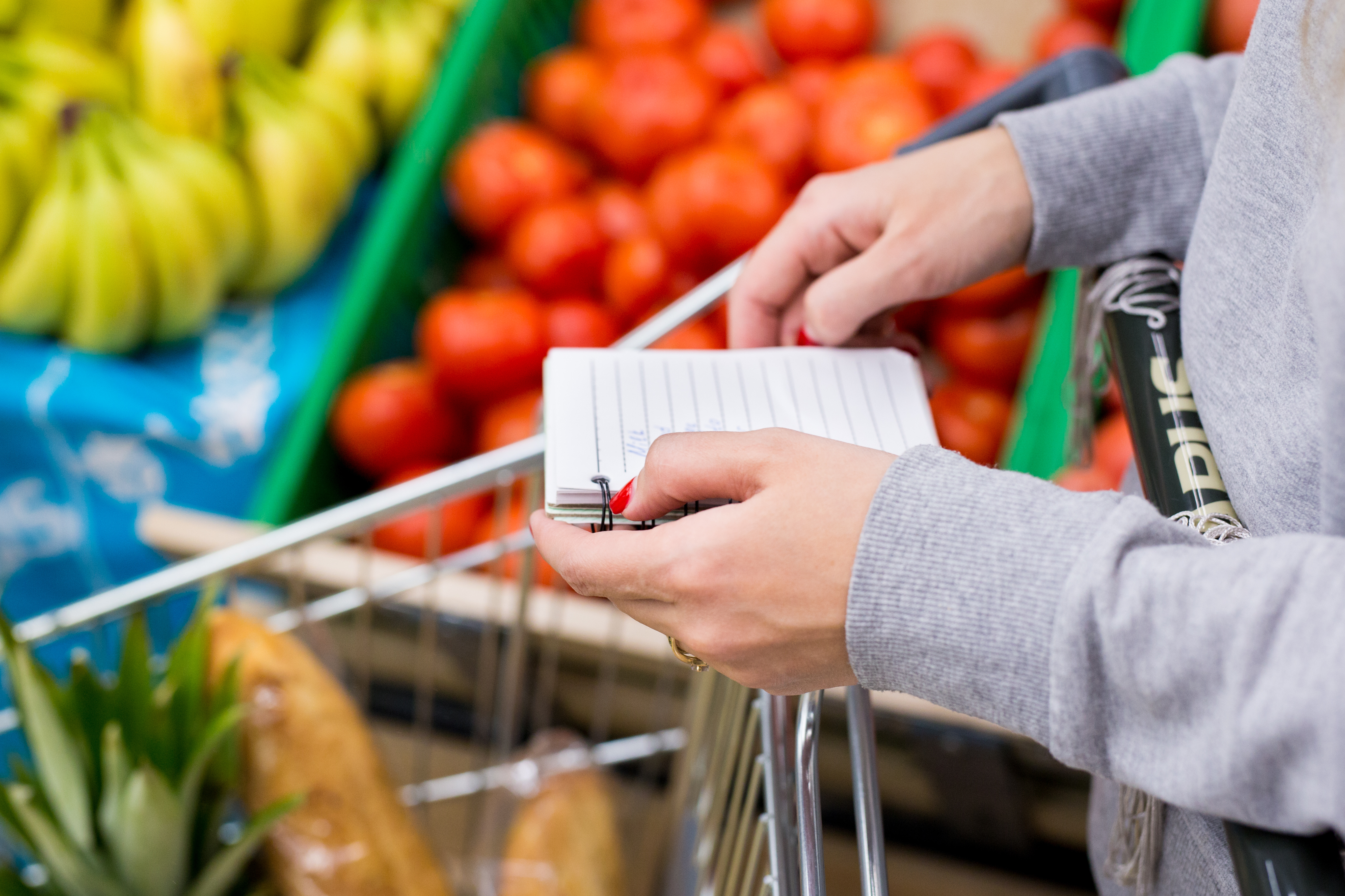 Person holding a shopping list while pushing a grocery cart filled with fresh produce in a supermarket.