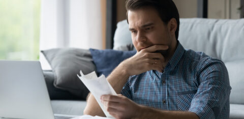 Man sitting on a couch, looking at bills with a concerned expression while using a laptop.