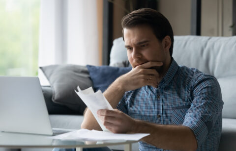 Man sitting on a couch, looking at bills with a concerned expression while using a laptop.