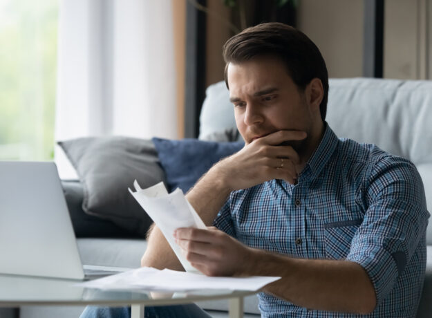Man sitting on a couch, looking at bills with a concerned expression while using a laptop.