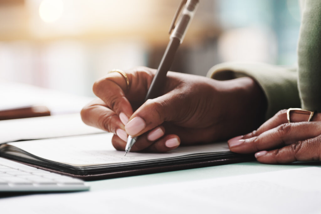 Close-up of a person writing in a notebook with a pen, surrounded by paperwork.