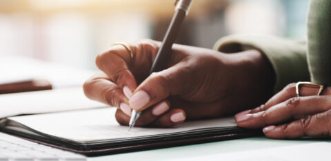 Close-up of a person writing in a notebook with a pen, surrounded by paperwork.