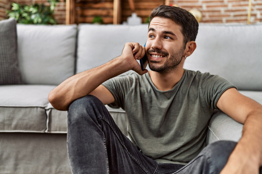 Young man sitting on the floor at home, smiling while talking on a smartphone.