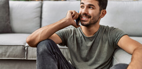 Young man sitting on the floor at home, smiling while talking on a smartphone.