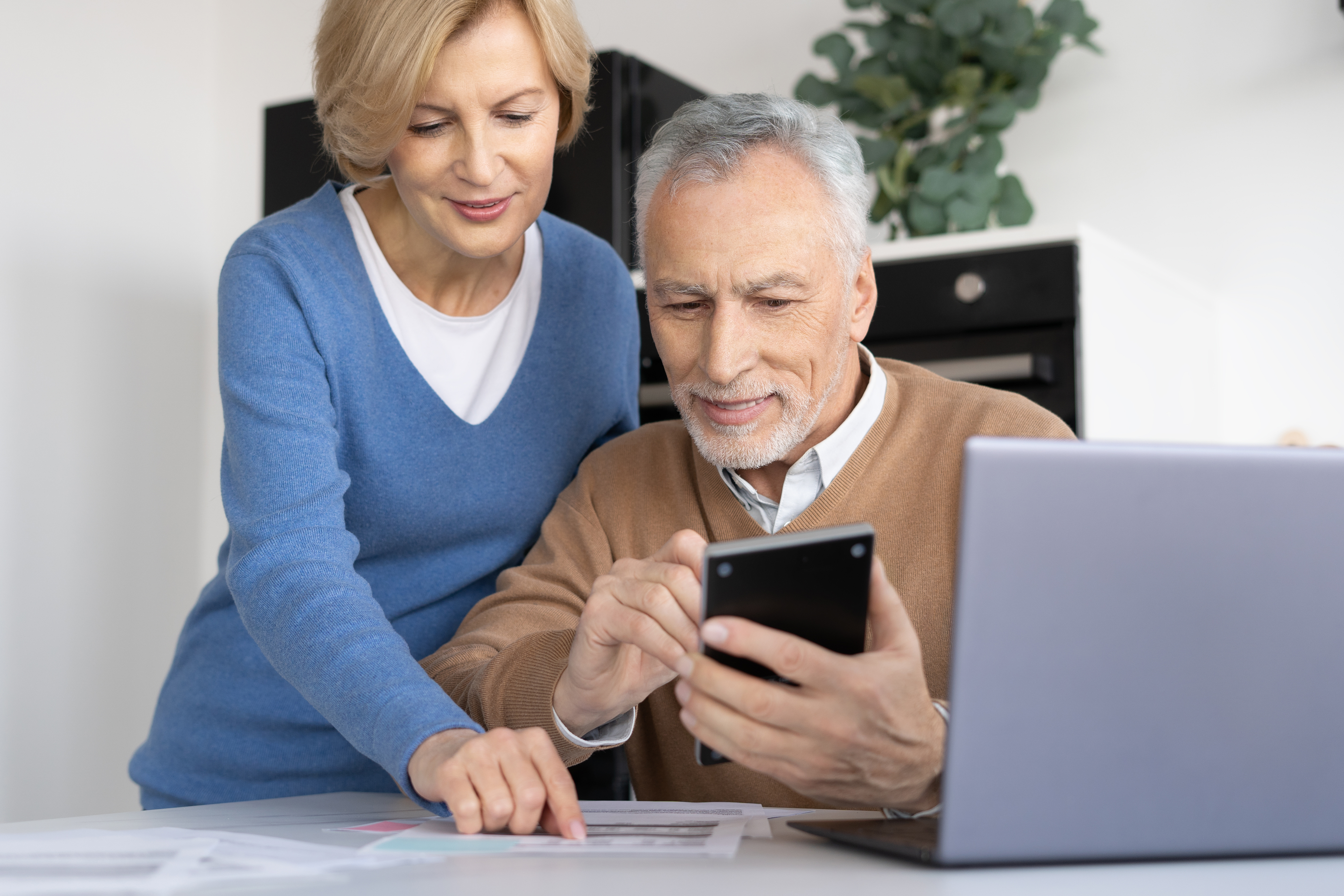 Older couple reviewing information on a smartphone, with papers and a laptop on the table.