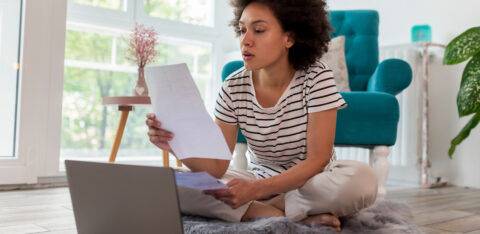 A woman sitting cross-legged on a rug, reviewing paperwork while using a laptop in a bright living room.
