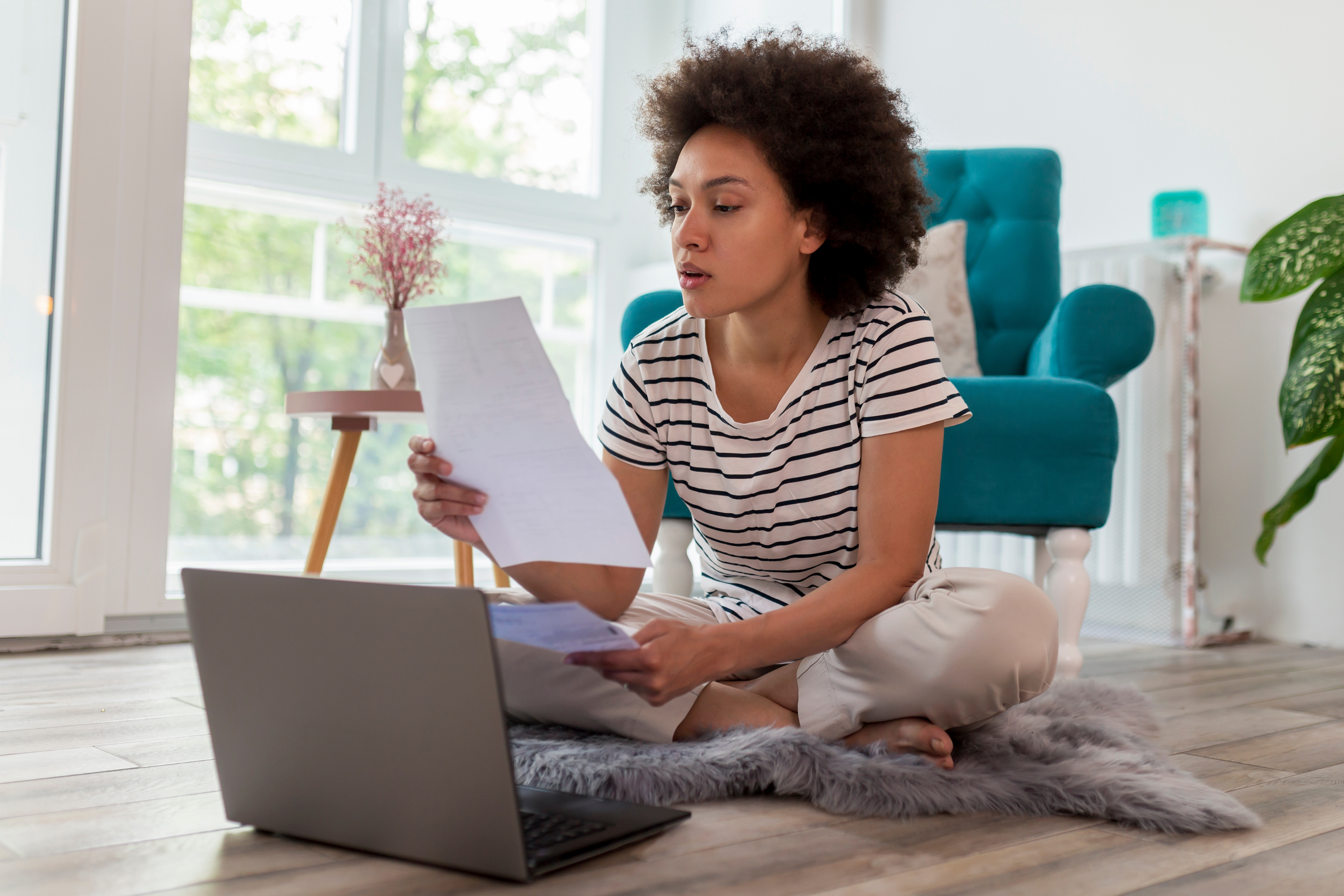 A woman sitting cross-legged on a rug, reviewing paperwork while using a laptop in a bright living room.