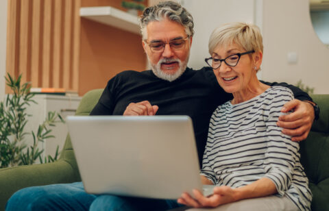 Smiling older couple sitting on a couch, looking at a laptop together.