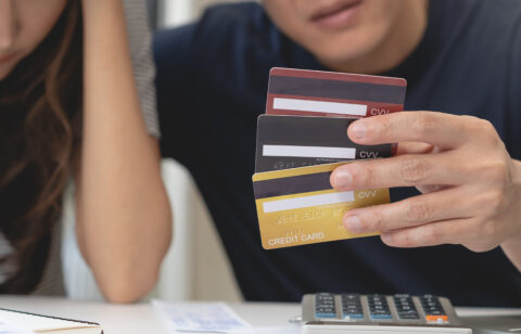 Person holding several credit cards while sitting at a table with a calculator and papers; another person appears stressed beside them.