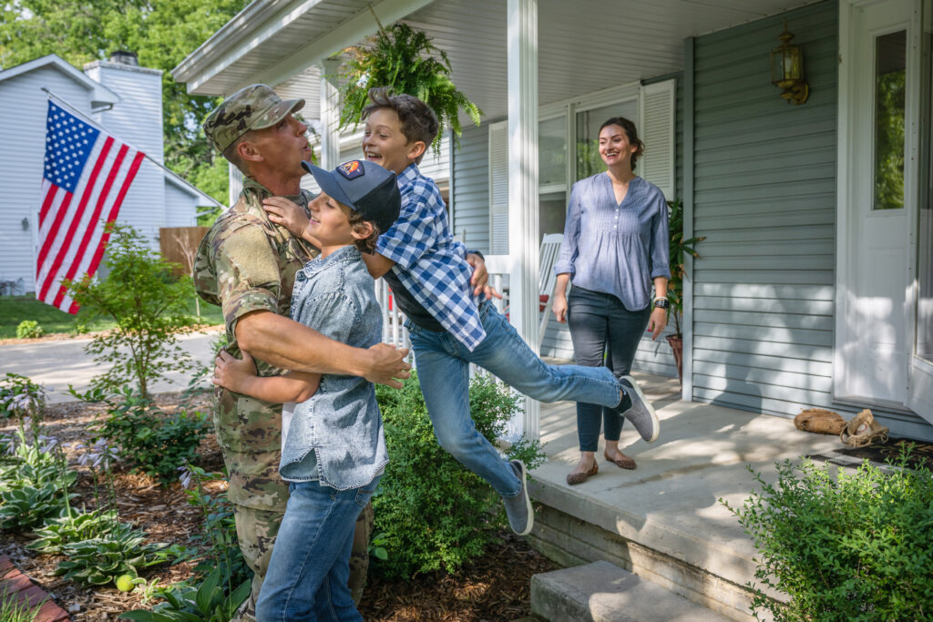 Military service member hugging two children in front of a house, with an American flag in the background and a smiling woman on the porch.