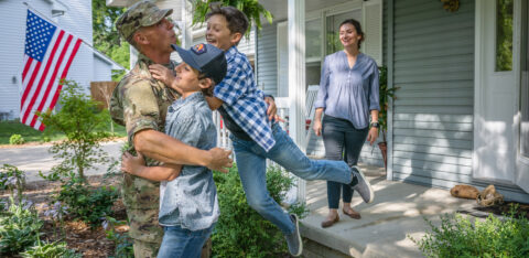 Military service member hugging two children in front of a house, with an American flag in the background and a smiling woman on the porch.