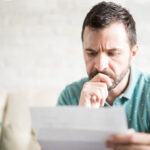 Man sitting on a couch, looking worried while reading a letter or bill.
