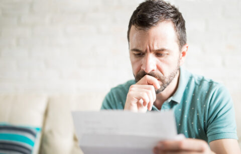 Man sitting on a couch, looking worried while reading a letter or bill.