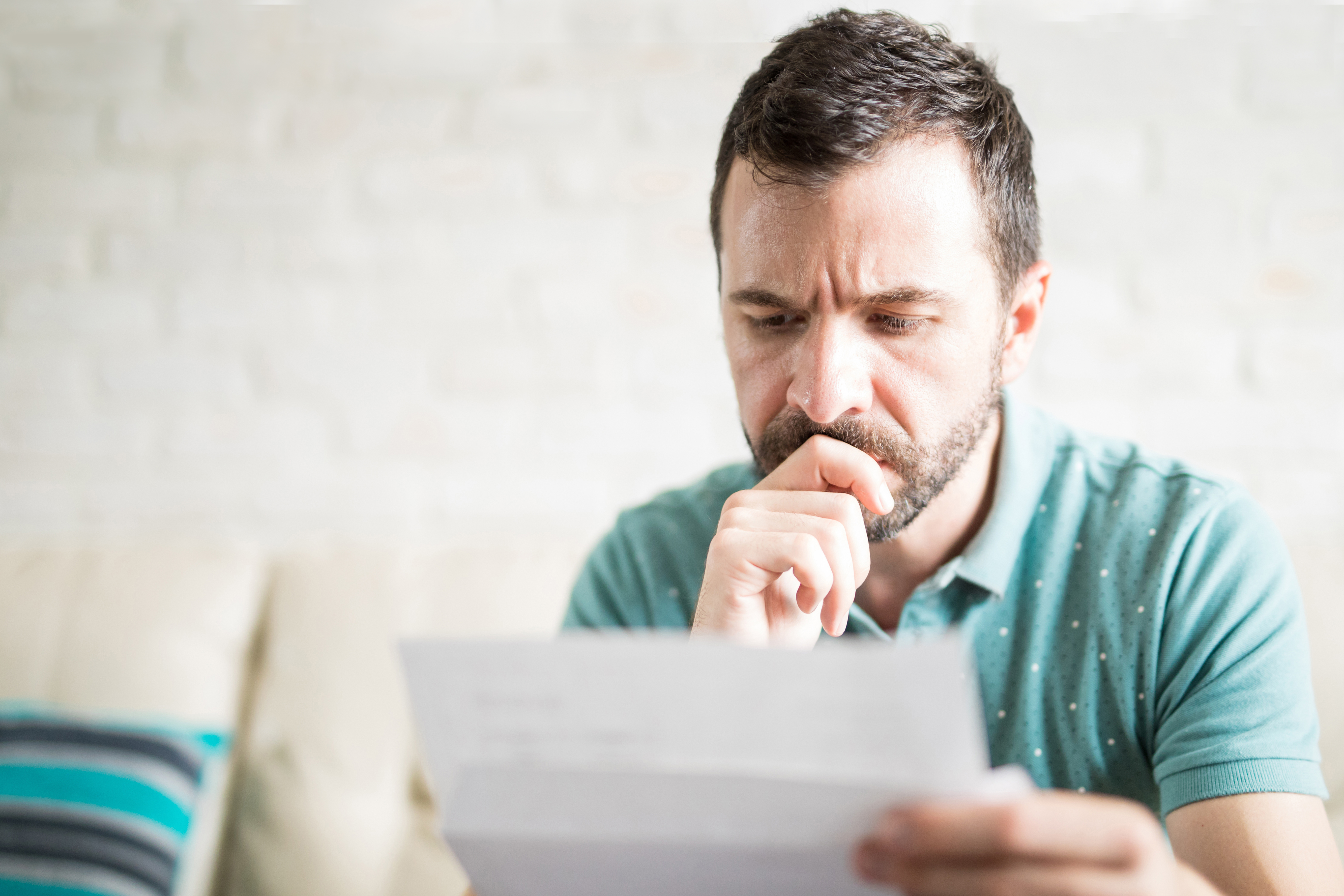 Man sitting on a couch, looking worried while reading a letter or bill.