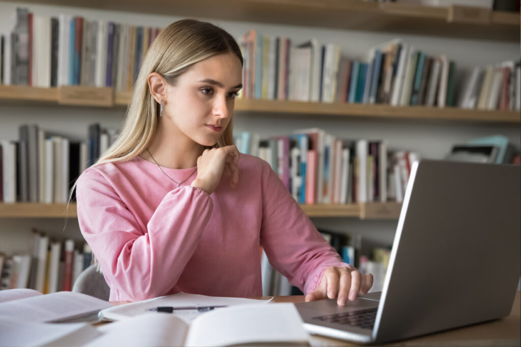 Focused young woman in a pink sweater studying at a desk with a laptop and open books.