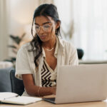 Woman working at a laptop while writing notes in a notebook at her desk.