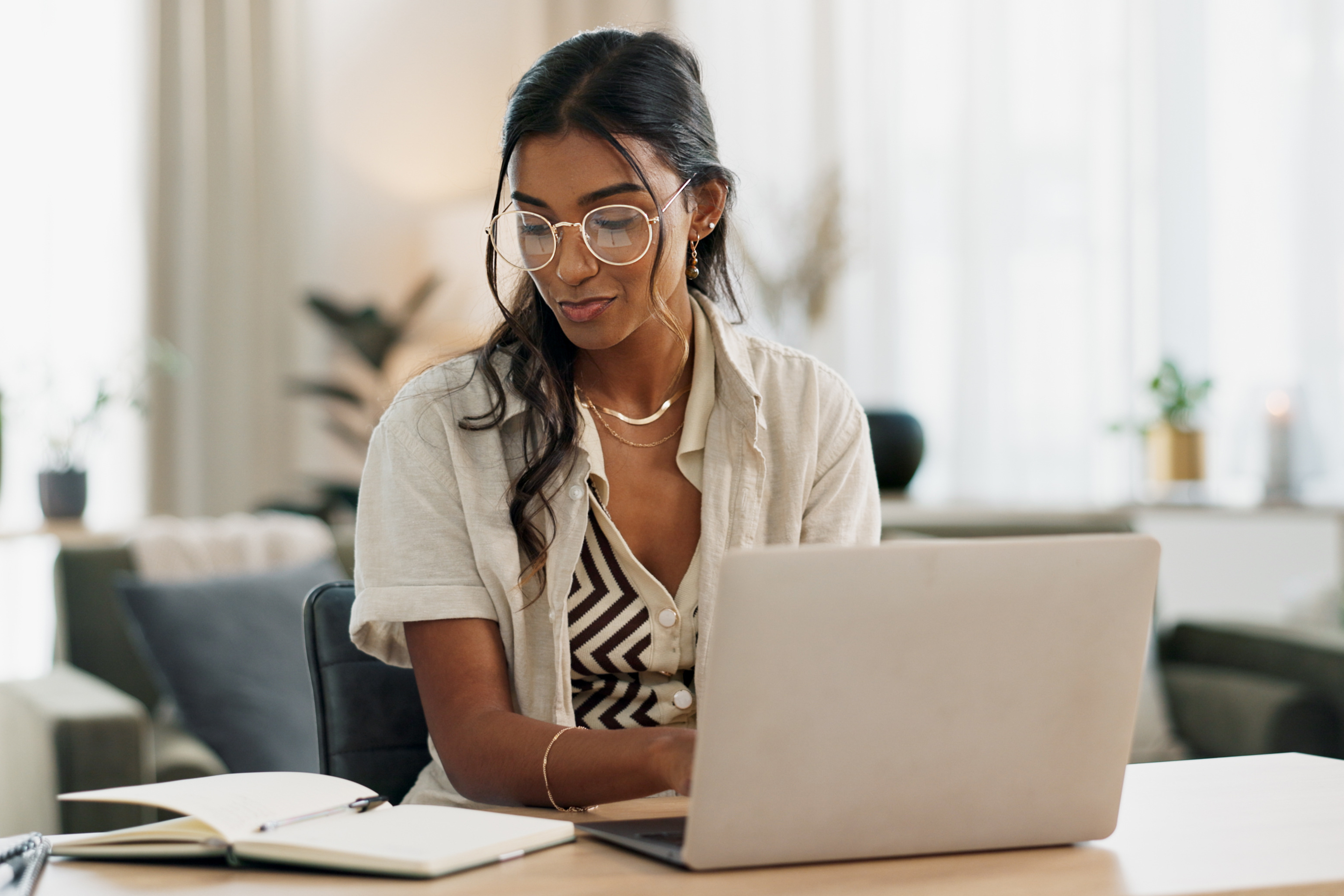 Woman working at a laptop while writing notes in a notebook at her desk.
