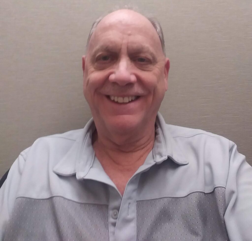 Smiling older man wearing a light gray collared shirt, seated in front of a neutral background.