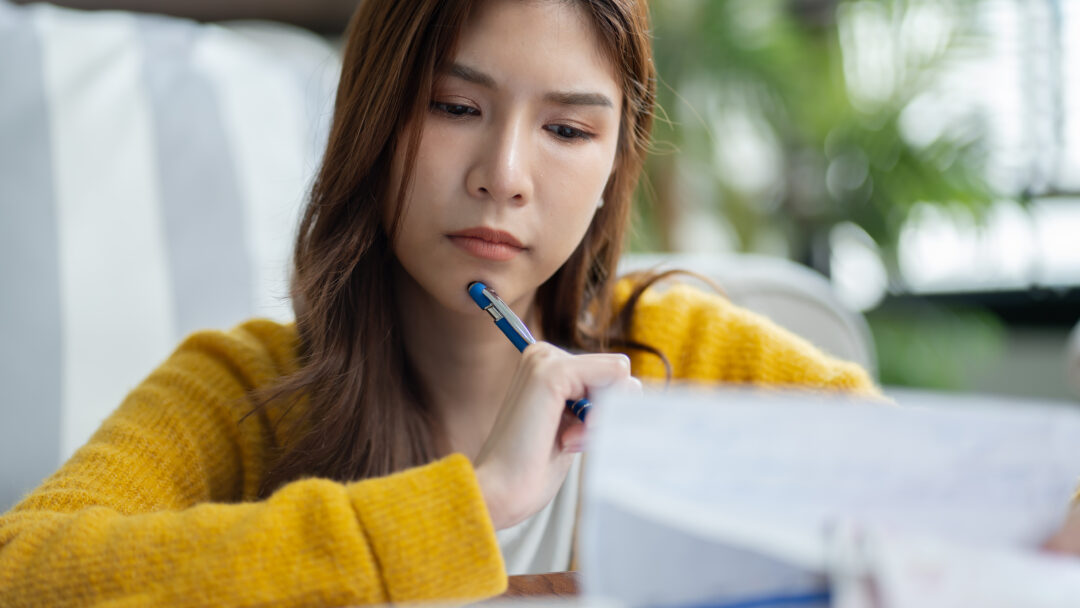 Young woman in a yellow sweater looking concerned while reviewing bills with a pen in hand.