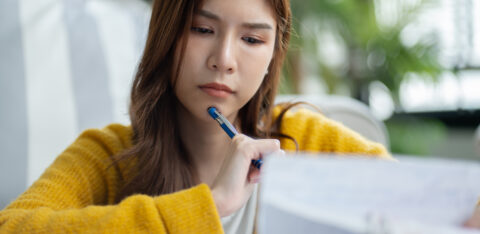 Young woman in a yellow sweater looking concerned while reviewing bills with a pen in hand.