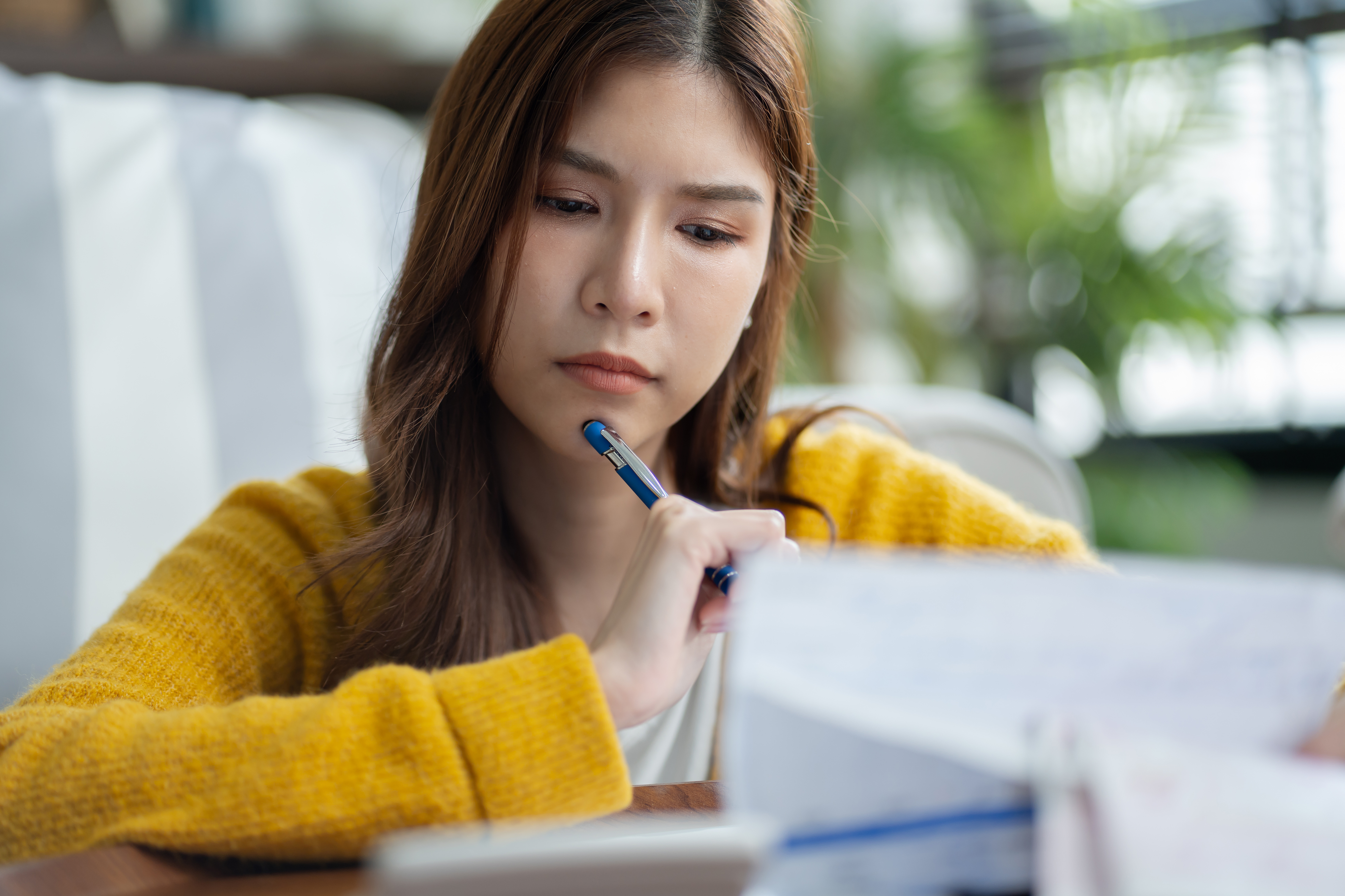 Young woman in a yellow sweater looking concerned while reviewing bills with a pen in hand.