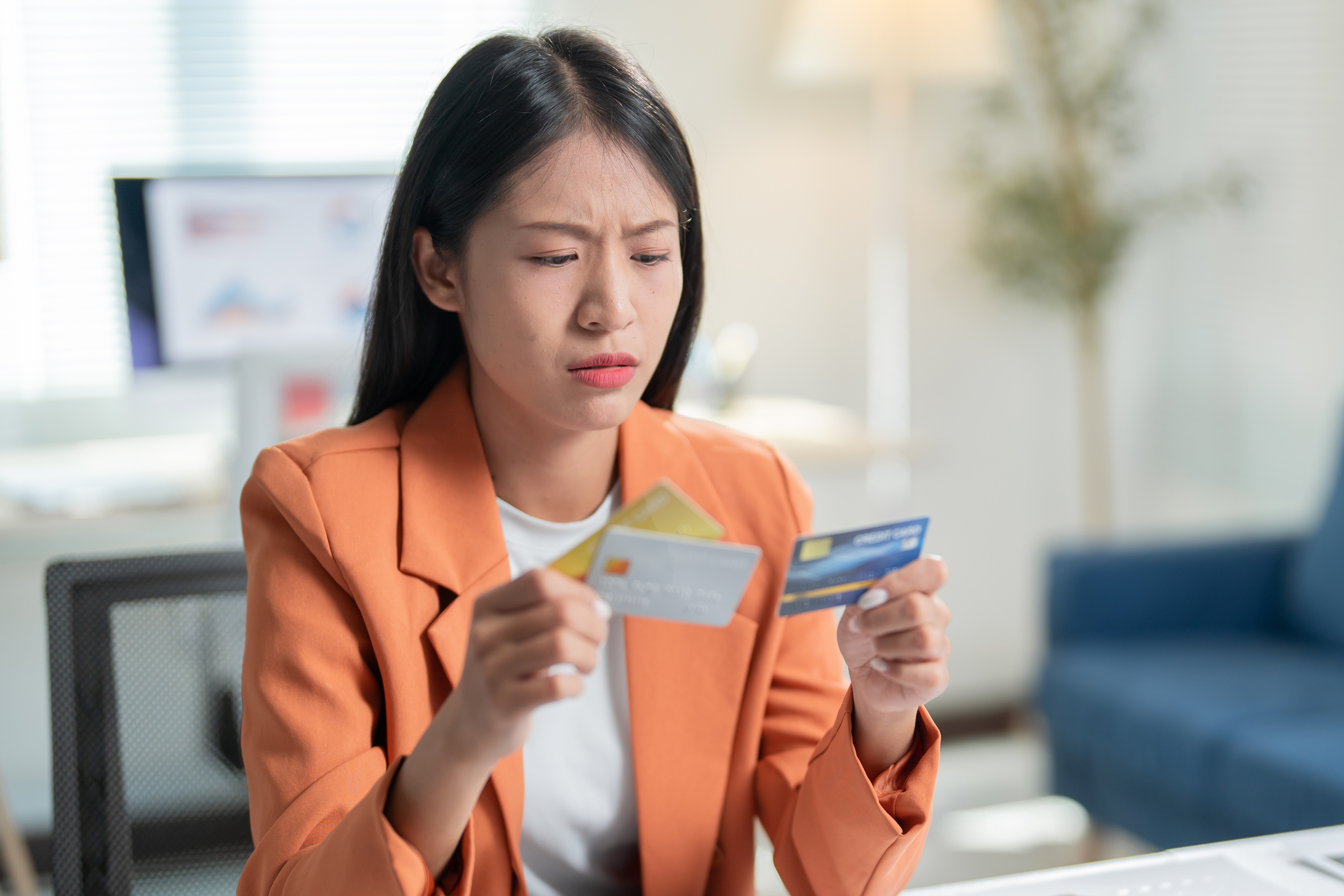 Young woman looking worried while holding multiple credit cards at her desk.