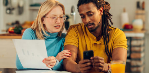 Couple sitting at a kitchen table reviewing financial documents together, with a phone and open notebook in front of them.