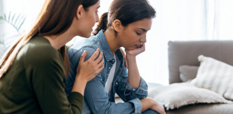 A woman sits with her head resting on her hand, looking upset, while another woman gently comforts her by placing a hand on her shoulder.