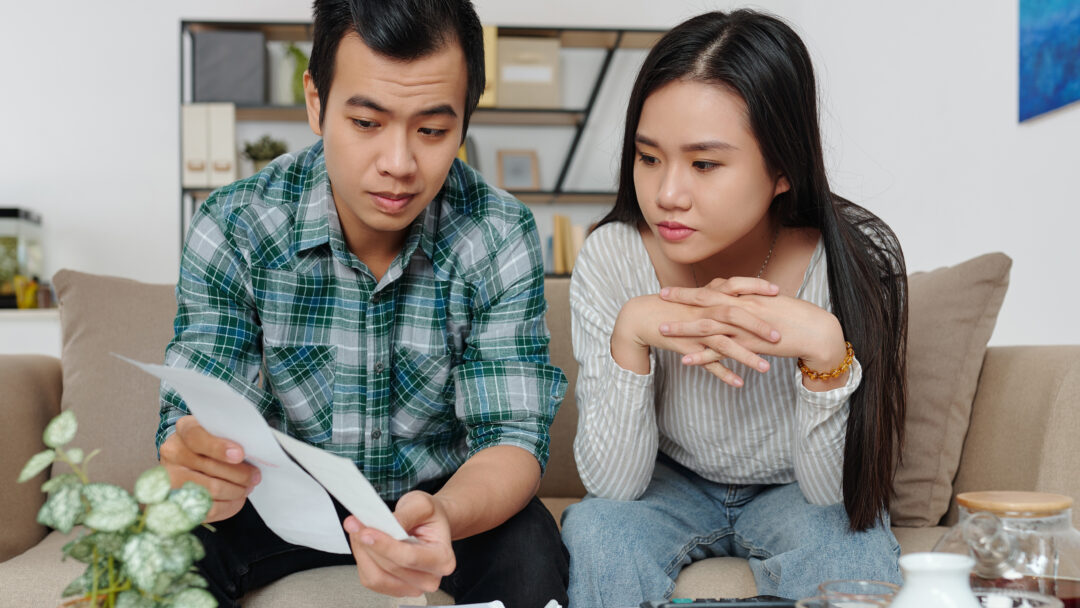 A couple sitting on the couch reviewing bills and financial documents with serious expressions.
