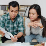 A couple sitting on the couch reviewing bills and financial documents with serious expressions.