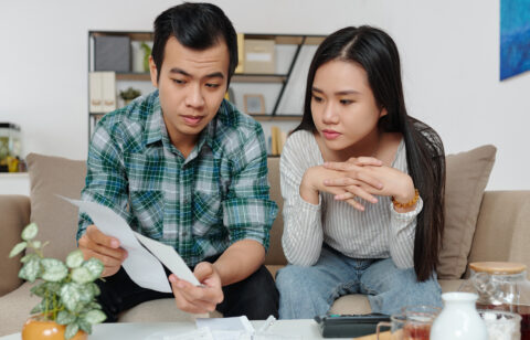 A couple sitting on the couch reviewing bills and financial documents with serious expressions.