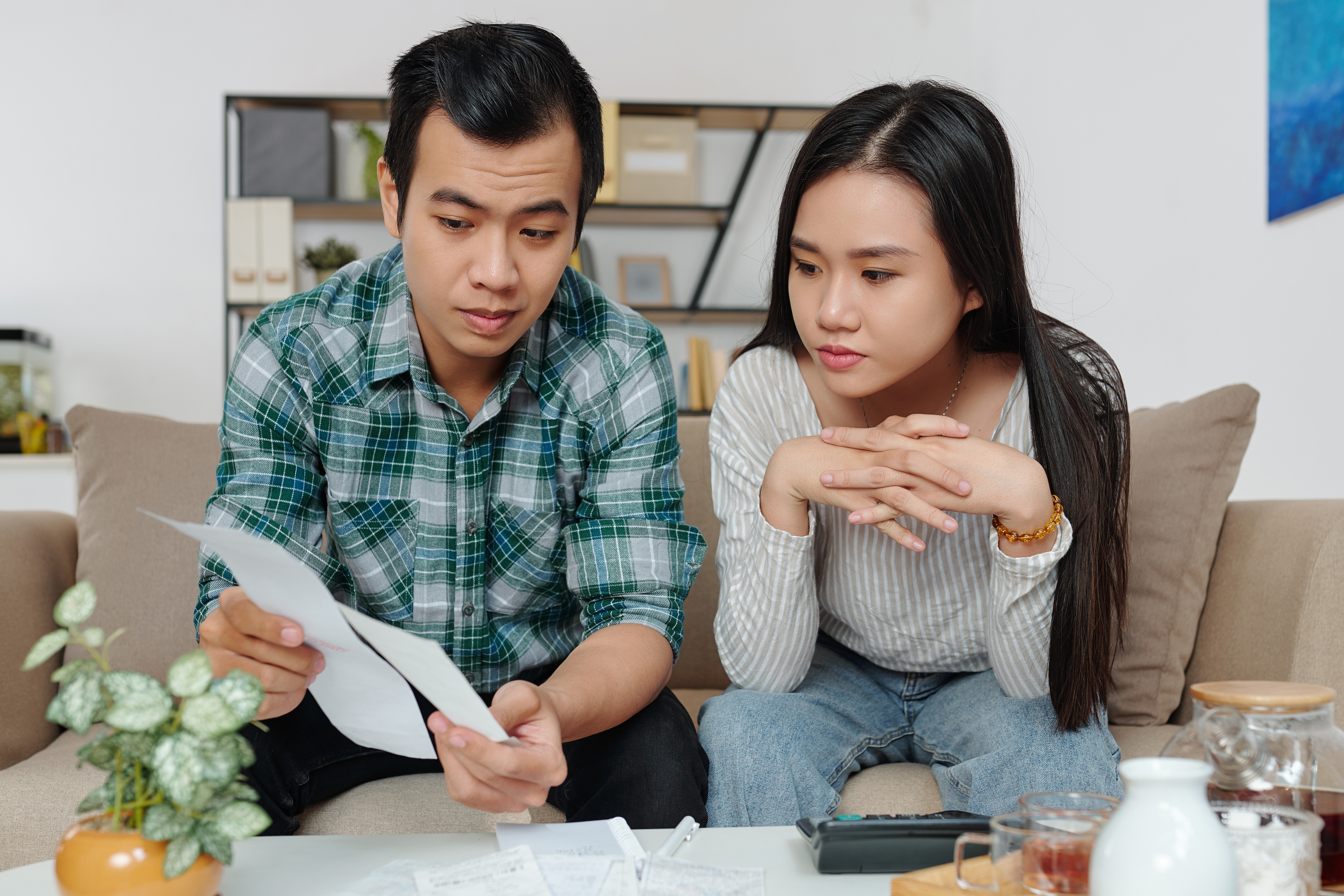 A couple sitting on the couch reviewing bills and financial documents with serious expressions.