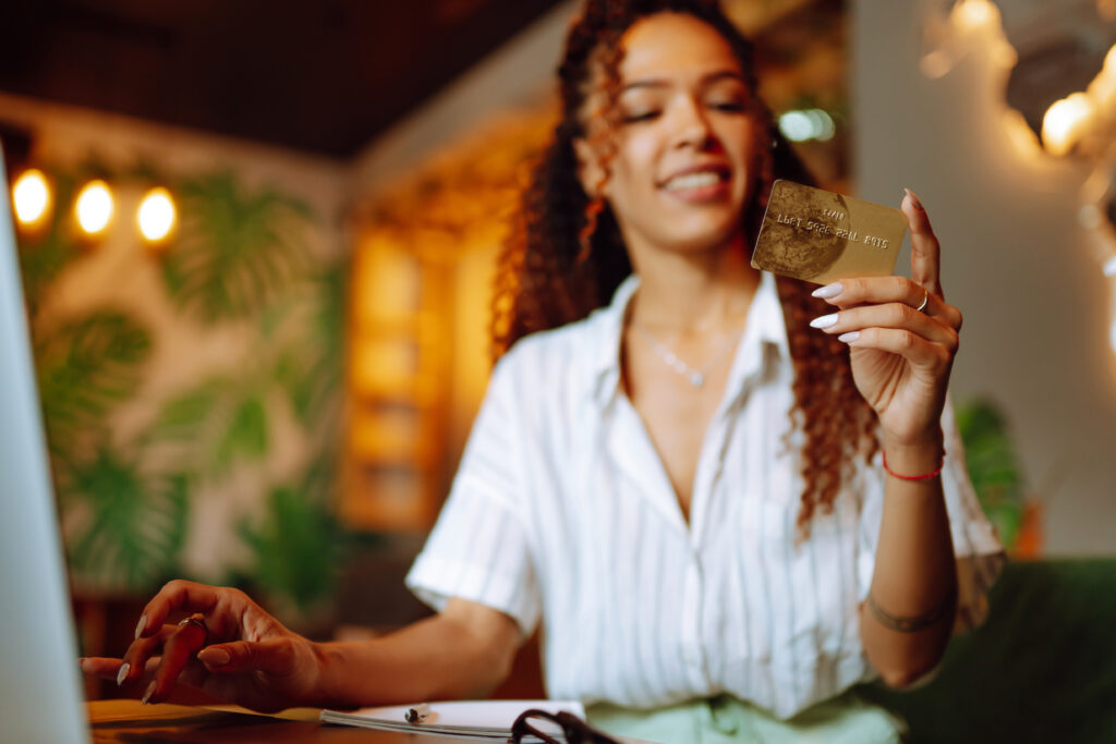 Smiling woman holding a gold credit card while sitting at a desk with a laptop.