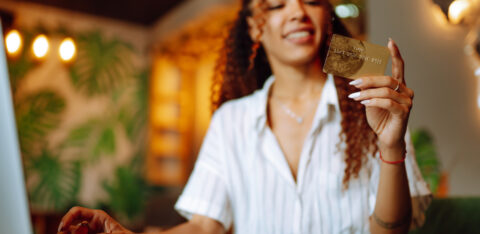 Smiling woman holding a gold credit card while sitting at a desk with a laptop.